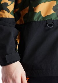 Kamouflagejacka med gröna och orange mönster, svart nedre del och kardborre- samt hårdvarudetaljer vid ärmsluten. Texturerad tyg.