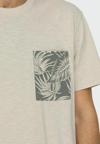 T-shirt grigio chiaro con taschino quadrato decorato da un motivo di foglie tropicali verde scuro. Tessuto morbido, scollatura a girocollo, maniche corte.