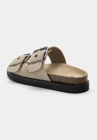 Beige Wildleder-Slipper mit zwei verstellbaren metallischen Schnallen, konturiertem Fußbett und einer schwarzen Gummisohle mit strukturiertem Grip.