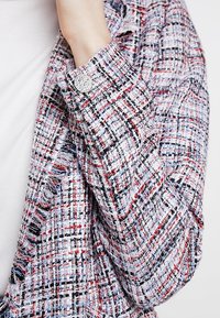 Gros plan d'une main ajustant le poignet d'une veste en tweed multicolore avec un bouton argenté sur un haut blanc.