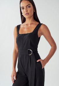 Žena s dlouhými tmavými vlasy, oblečená v černém bezrukávovém kombiné s čtvercovým výstřihem a páskem, stojící s jednou rukou v kapse na jednobarevném pozadí.