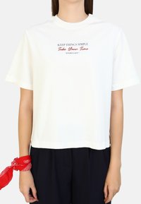 Hvid cropped T-shirt med korte ærmer. Har sort og rød trykt tekst: "HOLDF IN ENKELT Tag dig tid 'NYD LIVET'." Glat tekstur.