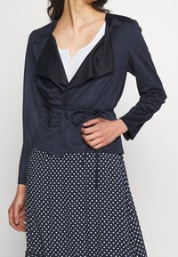 Donna che indossa un giacca avvolgente blu navy con revers neri sopra una maglietta bianca, abbinata a una gonna navy con pois, mentre sistema la cintura della giacca.