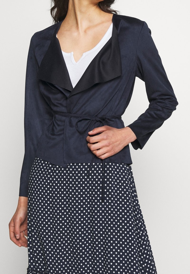 Donna che indossa un giacca avvolgente blu navy con revers neri sopra una maglietta bianca, abbinata a una gonna navy con pois, mentre sistema la cintura della giacca.
