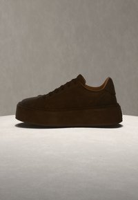 Σκούρο καφέ sneaker με πλατφόρμα και χοντρή σόλα, με κορδόνια μπροστά και λεία υφή, τοποθετημένο σε ανοιχτή επιφάνεια με ουδέτερο φόντο.