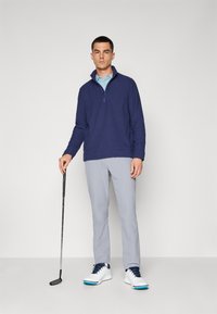 Pulôver com fecho em fleece azul-marinho, com gola alta, combinado com calças de golfe cinza claro e sapatos brancos com detalhes azuis. Taco de golfe na mão.