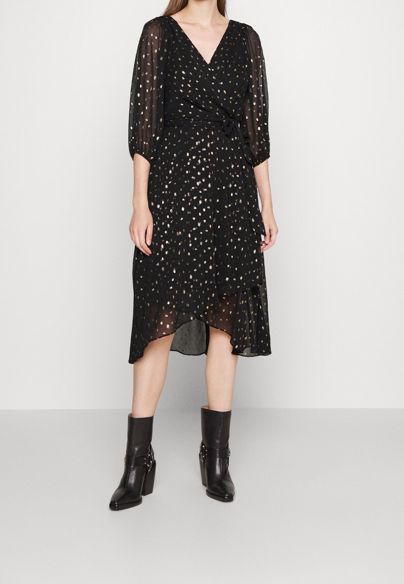 Robe noire à design portefeuille, manches 3/4 transparentes et motif tacheté doré. Mi-longue avec un ourlet à plusieurs couches, associée à des bottines noires.