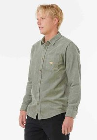 Grüne Cordhemd mit langen Ärmeln, einem Button-Down-Kragen und einer aufgesetzten Tasche mit einem rautenförmigen, beige Akzent.