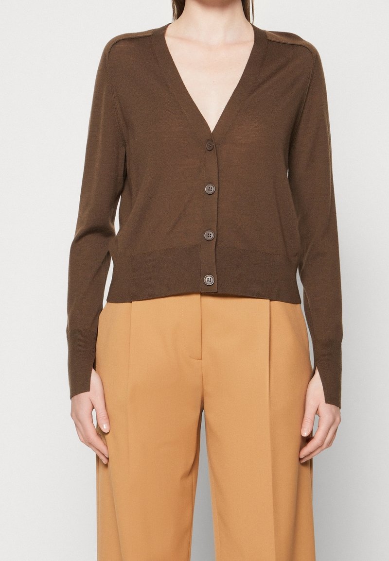 Cardigan en tricot marron avec un col en V, des boutons sur le devant et des manches longues, associé à un pantalon beige taille haute. Texture douce et design ajusté.