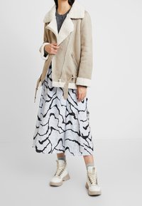 Beige suede jacket with cream shearling collar and cuffs, zip closure, and side pockets, paired with a light blue skirt featuring black abstract patterns and white high-top boots.