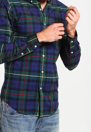 Homme portant une chemise à carreaux bleus, verts et rouges boutonnée, attachant le poignet de sa manche droite, portant un jean bleu clair.