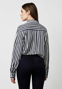 Striped blouse in navy and white, featuring long sleeves, a buttoned cuff, and a curved hem, paired with black fitted pants.