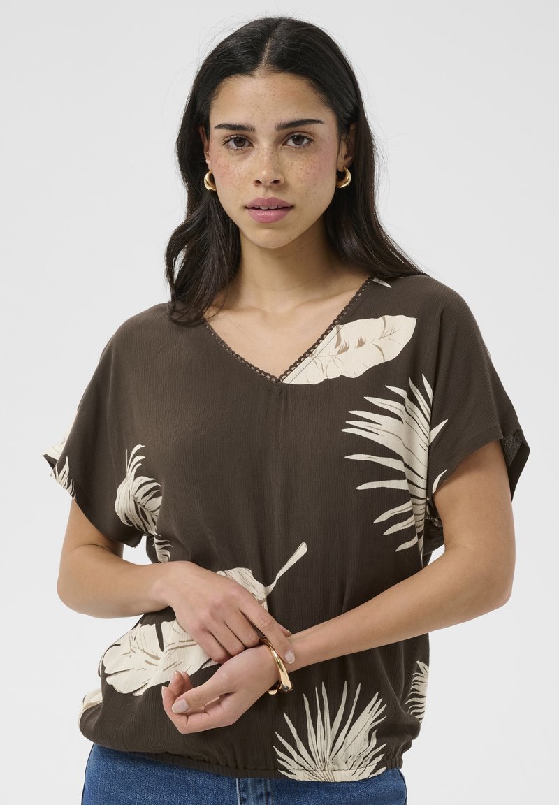 Jeune femme aux cheveux foncés portant des boucles d'oreilles créoles dorées, un haut marron avec des imprimés de feuilles tropicales beiges et un jean bleu, tenant son poignet.