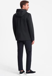 Schwarze Kapuzenjacke mit sleekem Design und glatter Textur, kombiniert mit dunklen Hosen und schwarzen Schuhen, von hinten betrachtet.