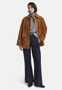 Giacca in suede marrone con grandi tasche, abbinata a un maglione grigio a dolcevita e jeans in denim scuro a gamba larga con cuciture a contrasto.