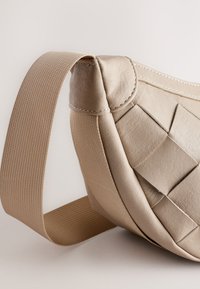 Beige läderväska med en strukturerad vävd design på sidan och ett brett, slätt tygrem. Har kontrasterande sömdetaljer.