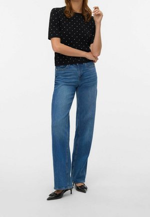 Vrouw die hoge taille rechte blauwe jeans draagt, een zwart polkadot kortärmelig topje en zwarte spitse kittenheels, poseert met gekruiste armen.