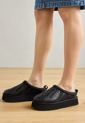 Jambes portant des chaussures plateformes noires sans lacets avec des motifs cousus, associées à une jupe en jean longueur genou sur un sol en bois.