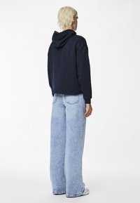 Felpa blu navy con cappuccio regolabile, abbinata a jeans a gamba larga di colore azzurro chiaro. Il tessuto sembra morbido, con una vestibilità rilassata e dettagli minimi.