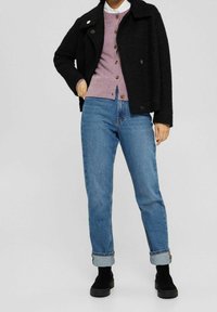 Person som bär en svart fluffig jacka, en lavendelfärgad knäppt cardigan över en vit skjorta, uppvikta blå jeans och svarta platåstövlar som står mot en grå bakgrund.