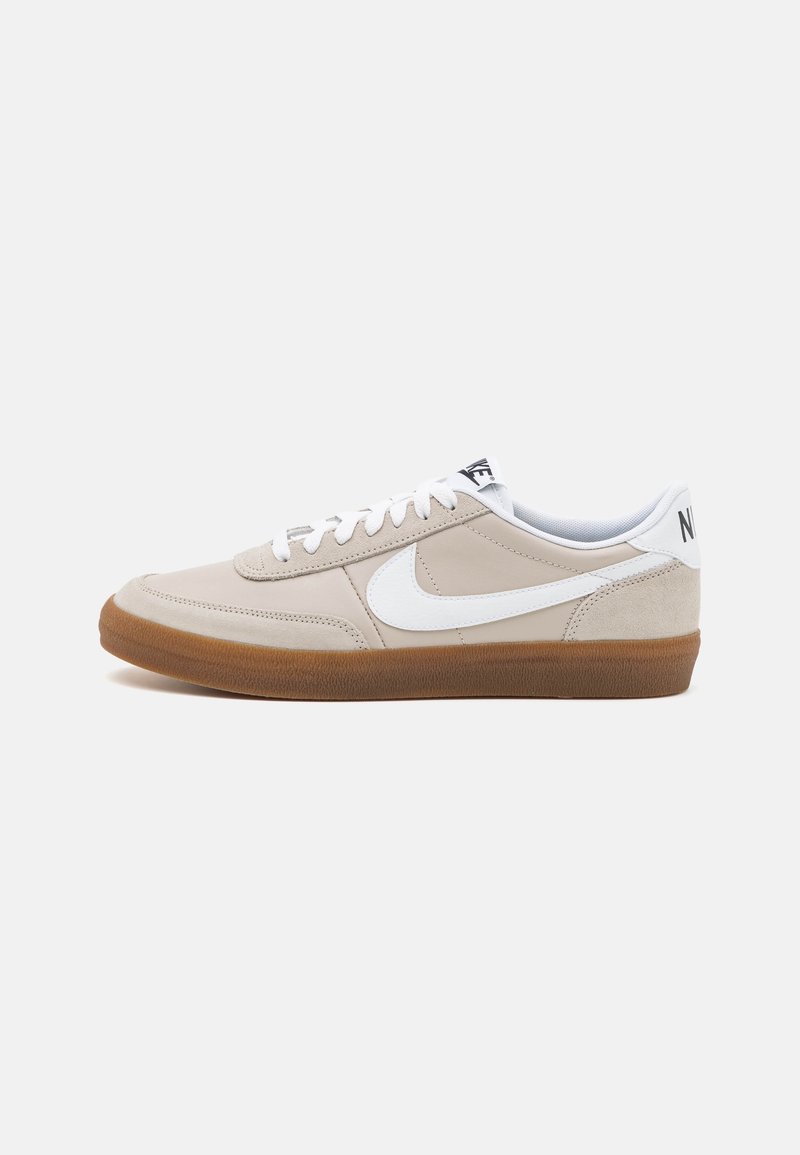 Beige urheilujalkine valkoisilla yksityiskohdilla, jossa on teksturoitu päällinen, litteät nauhat ja kumipohja. Logo kantapäässä ja sivussa.
