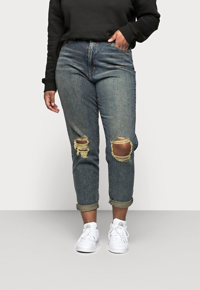 Niebieskie dżinsy z przetarciami, z dwoma dziurami na kolanach i zrolowanymi mankietami, w połączeniu z białymi trampkami i czarną bluzą.