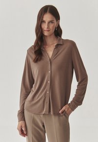 Camicia marrone a maniche lunghe con colletto, leggero lucido, chiusura con bottoni e morbida texture, abbinata a pantaloni beige.