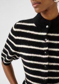 Melnā un baltā svītraina kardigāna, kas ir ar plašu apkakli, īsām piedurknēm un pogu priekšējo aizdari ar apaļām melnām pogām. Tekstūrēta adījuma materiāls.