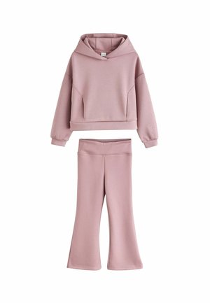 Ung pige med håret i en knold iført en pink hættetrøje og leggings, som holder en hvid hulahopring, smilende og kigger til siden.