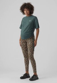 Umstandst-Shirt in Dunkelgrün mit Herzmotiv, kombiniert mit Leggings im Leopardenmuster und schwarzen Plateau-Sneakern. Glatter Stoff, lockere Passform.