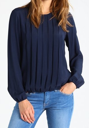 Frau trägt eine marineblaue Bluse mit langen Ärmeln und vertikalen Falten, kombiniert mit hellblauen Jeans, eine Hand in der Tasche und die andere auf dem Oberschenkel ruhend.