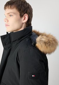 Parka neagră cu o glugă căptușită cu blană. Prezintă un închidere cu fermoar și capse, țesătură texturată și un ecuson cu brandingul "Tommy Hilfiger".