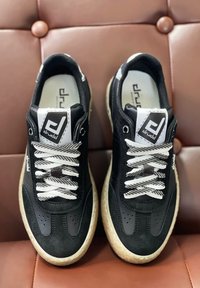 Zapatilla negra con una combinación de texturas lisas y de ante, con cordones a rayas blancas y negras, suela de goma y logo en la lengüeta.
