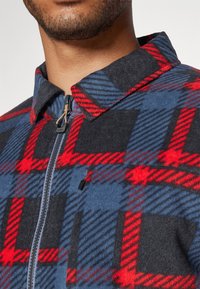 Kockovaný fleecov�ý kabát so vzormi v červenej, modrej a čiernej farbe. Má zips a golier s textilným akcentom pri zipsi.