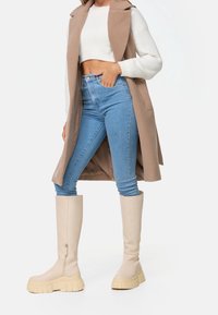 Femme portant un long gilet beige, un pull court blanc, un jean skinny taille haute bleu clair, et des bottes montantes à plateforme épaisses couleur crème.