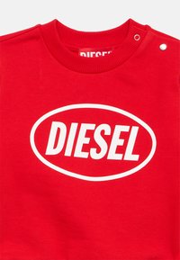 Rød collegegenser laget av bomull, med en stor hvit oval hvor "DIESEL" er trykt med fete bokstaver. Har trykknappdetaljer på skulderen.