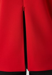 Vêtement rouge à texture lisse, doté d'une fermeture éclair au dos et d'une fente à l'ourlet, complété par un tissu de fond noir.