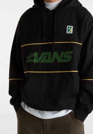 Sweat à capuche noir Vans avec logo vert et rayures jaunes, porté sur une chemise blanche, associé à un pantalon marron, mains dans les poches.