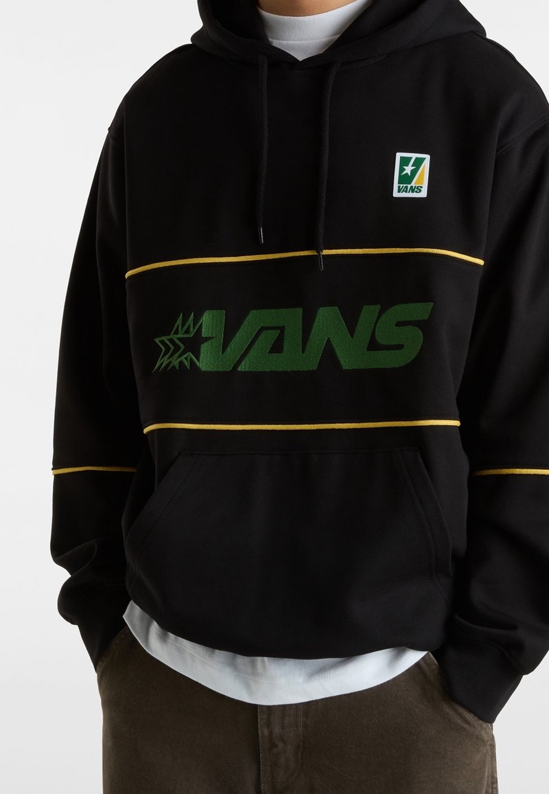 Sweat à capuche noir Vans avec logo vert et rayures jaunes, porté sur une chemise blanche, associé à un pantalon marron, mains dans les poches.