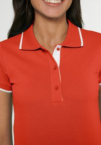 Rode polo met een gestructureerde stof, twee knopen en witte accenten op de kraag en mouwen. Close-up van de halslijn.