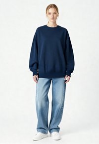 Felpa oversize blu navy in tessuto morbido, con scollo rotondo e polsini a costine, abbinata a jeans a gamba larga azzurri e sneakers bianche.