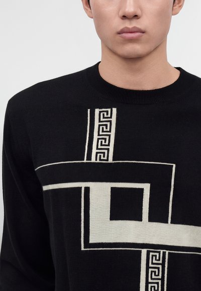 Jeune personne portant un pull noir avec un carré géométrique blanc et un motif clé grecque sur le devant, visible des épaules au menton.