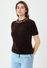 Brauner, flauschiger Kurzarm-Pullover mit einem juwelenbesetzten Ausschnitt. Einfaches Design mit weicher Textur, kombiniert mit hellblauen Jeans.