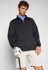 Svart zip-up genser med høy krage, brukt over en lys blå polo, kombinert med beige shorts og en hvit golfhanske som holder en kølle.