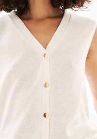 Vit ärmlös kofta med V-ringning, med två guldknappar framtill. Materialet har ett strukturerat stickat mönster.