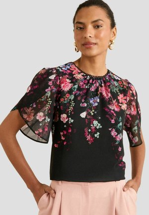 Frau in bluse mit schwarzen Blumenmotiven und durchsichtigen kurzen Ärmeln, in hellrosa Hose, posiert mit Händen in den Taschen vor einem einfarbigen grauen Hintergrund.