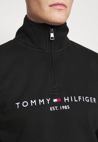 Uomo che indossa una felpa nera con zip, con il logo Tommy Hilfiger e l'emblema della bandiera sul petto, con la zip tirata fino al collo.