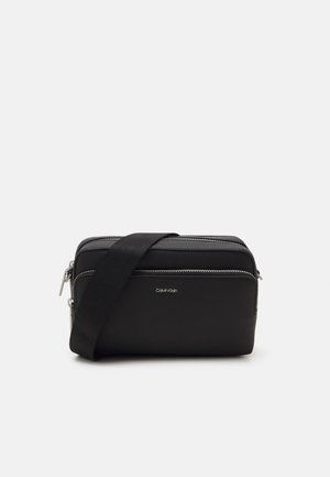 Zwarte Calvin Klein crossbodytas met twee ritssluitingen en een verstelbare schouderriem op een effen achtergrond.