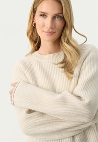 Cremefarvet sweater med struktureret design, brede ærmer og en rund halsudskæring, der fremviser et blødt og hyggeligt udseende.