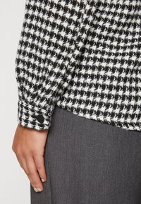 Blusa in bianco e nero con texture e motivo a scacchi. Presenta un polsino con un bottone nero. Indossata con un tessuto grigio.